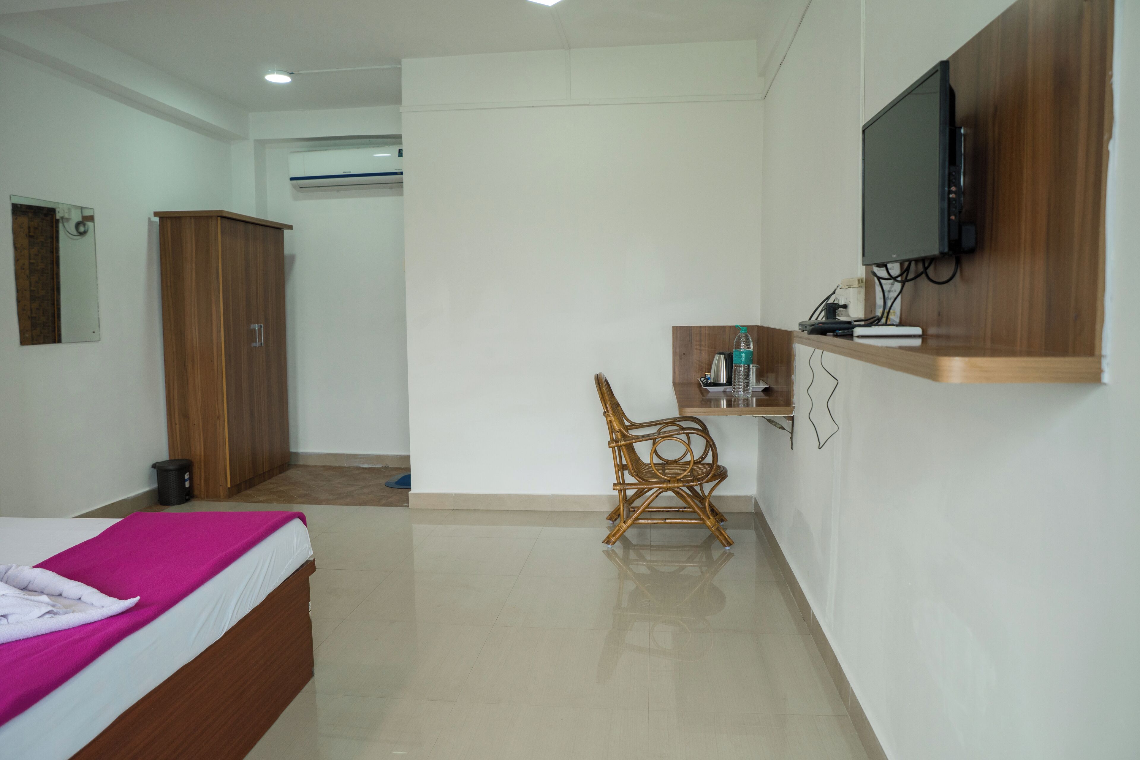 A hotel room with a bed, a wooden wardrobe, a television mounted on the wall, and a small wooden table with a chair.