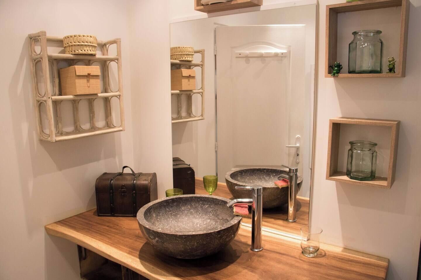 A bathroom with a large stone basin sink, a mirror, and shelves with baskets and jars.