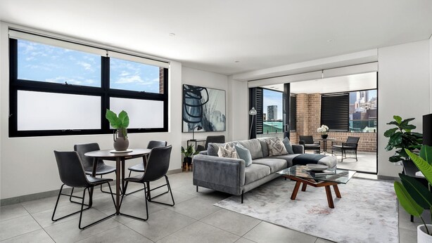 A modern living room with a grey sofa, a coffee table, and a dining area with chairs and a table. There are large windows with blinds, a potted plant, and artwork on the walls.