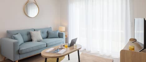 A modern living room with a light blue sofa, a wooden coffee table, a laptop, a glass of orange juice, and a television.