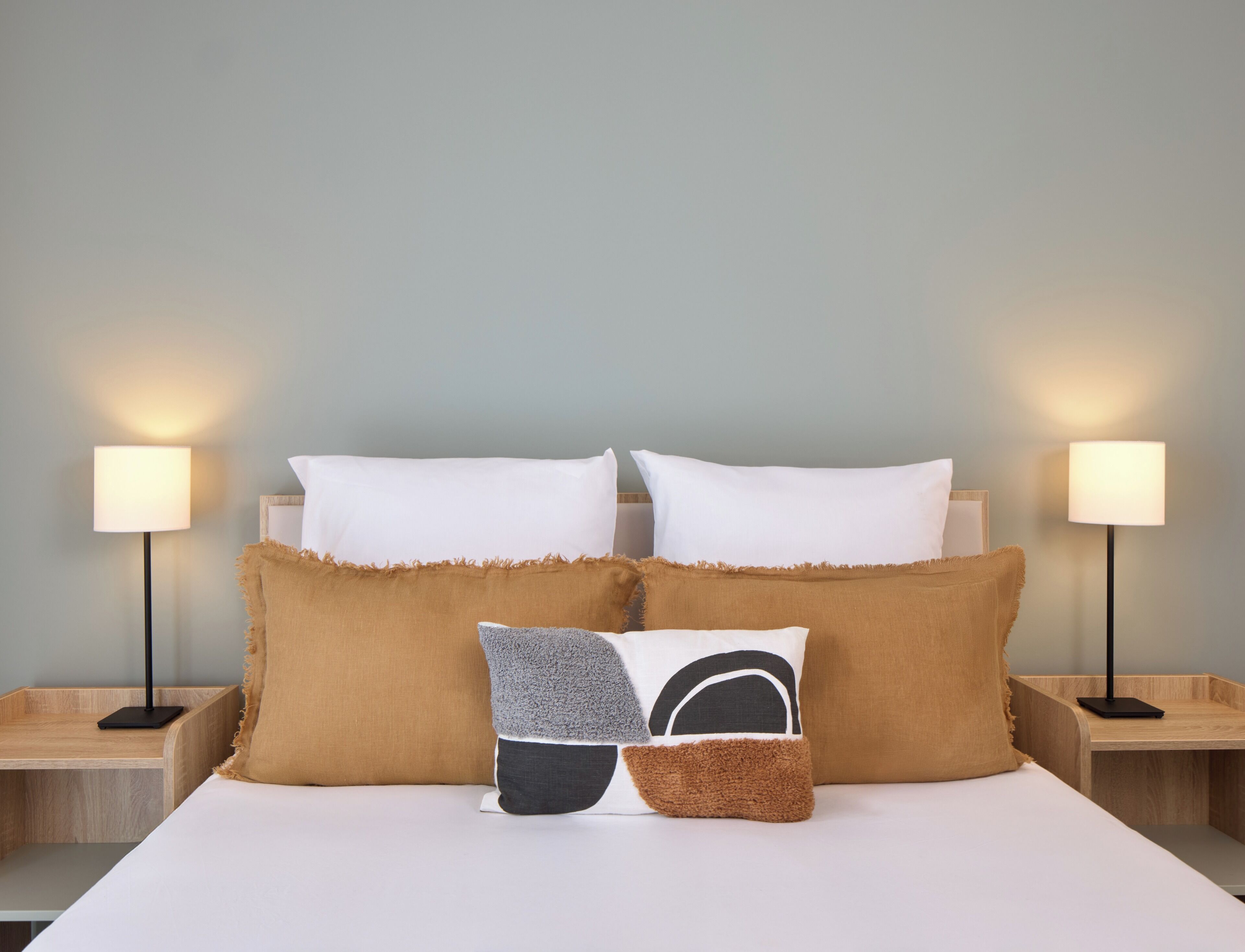 Een netjes opgemaakt bed met witte lakens en twee bedlampjes, een houten nachtkastje en een muur in een lichtblauwe kleur.