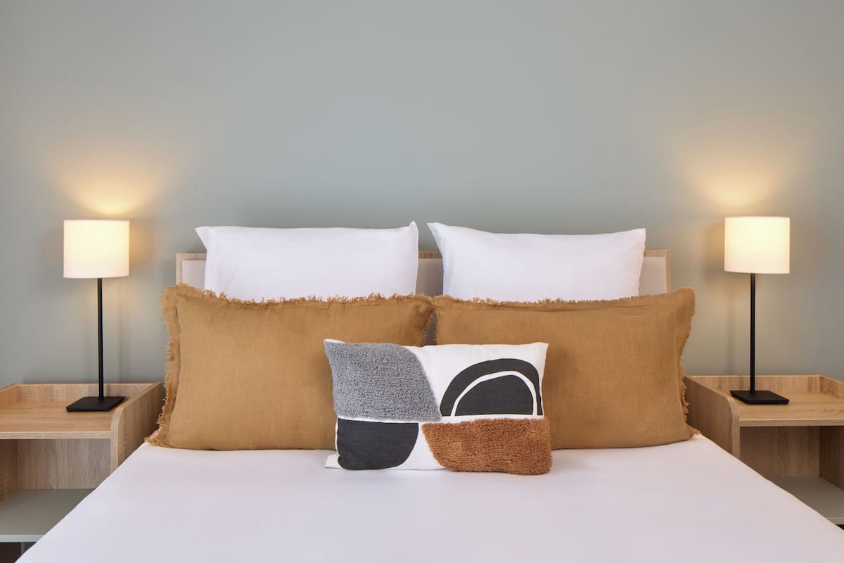 A neatly made bed with white bedding and two bedside lamps, a wooden nightstand, and a wall with a light blue color.