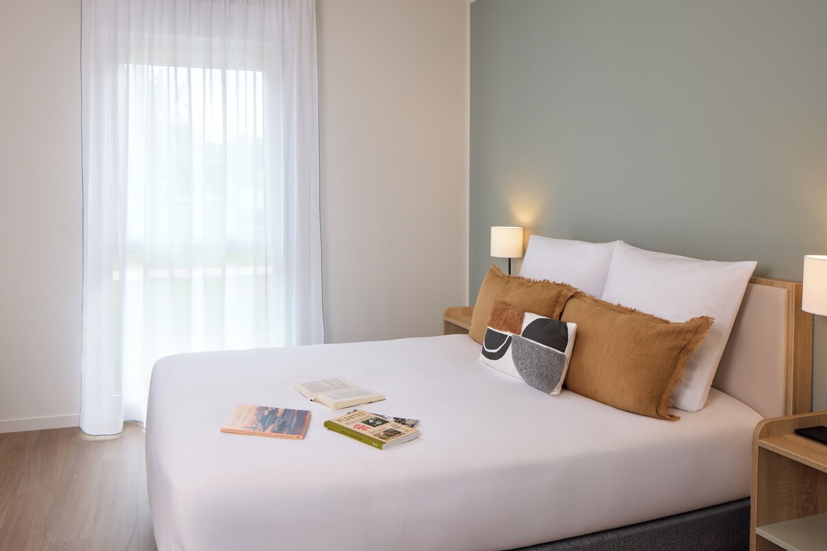 Een net opgemaakt bed met kussens, een bedlampje en boeken op een houten vloer.