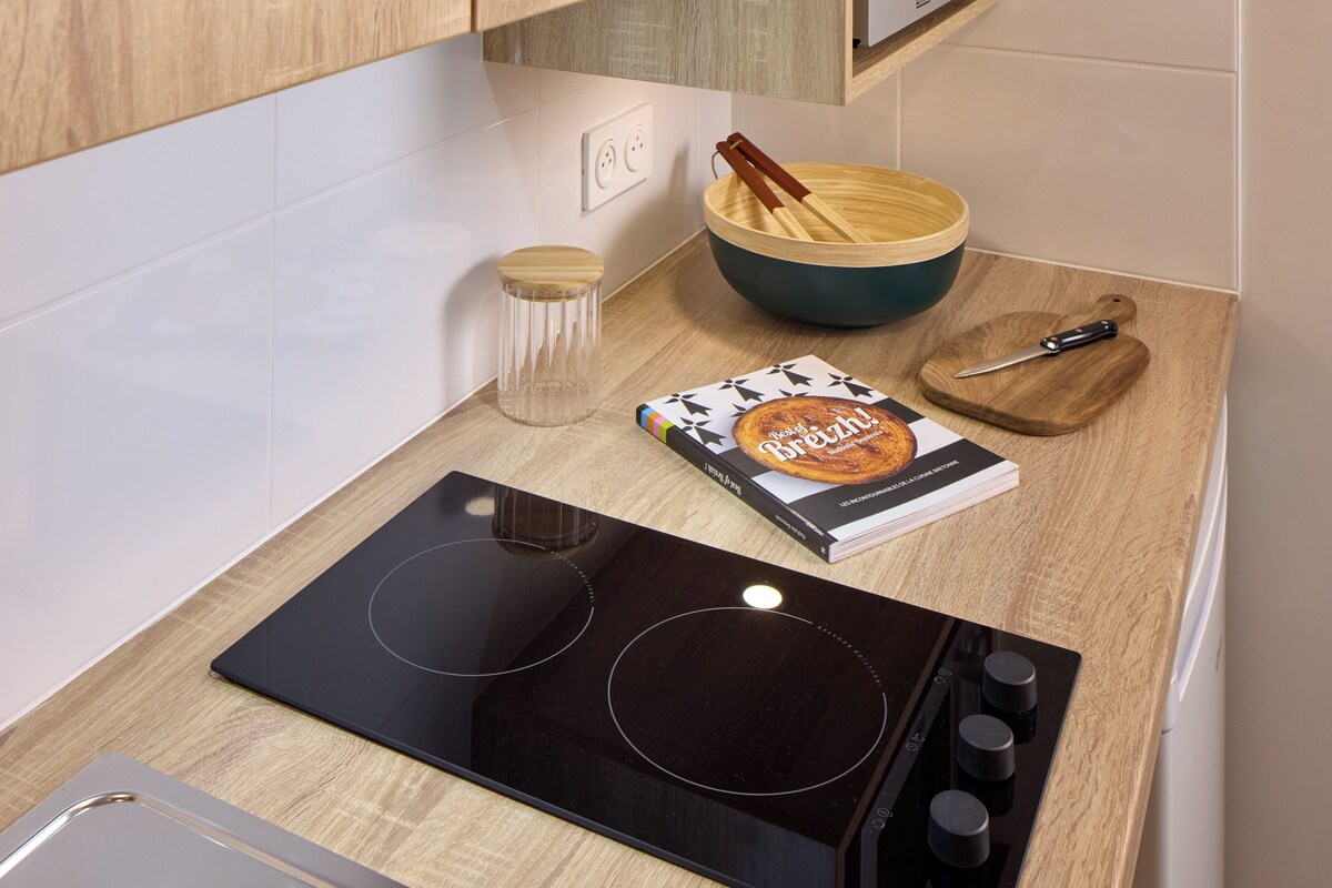 A kitchen with a black induction cooktop, wooden cabinets, a microwave, a wooden bowl, a book, and a wooden cutting board.