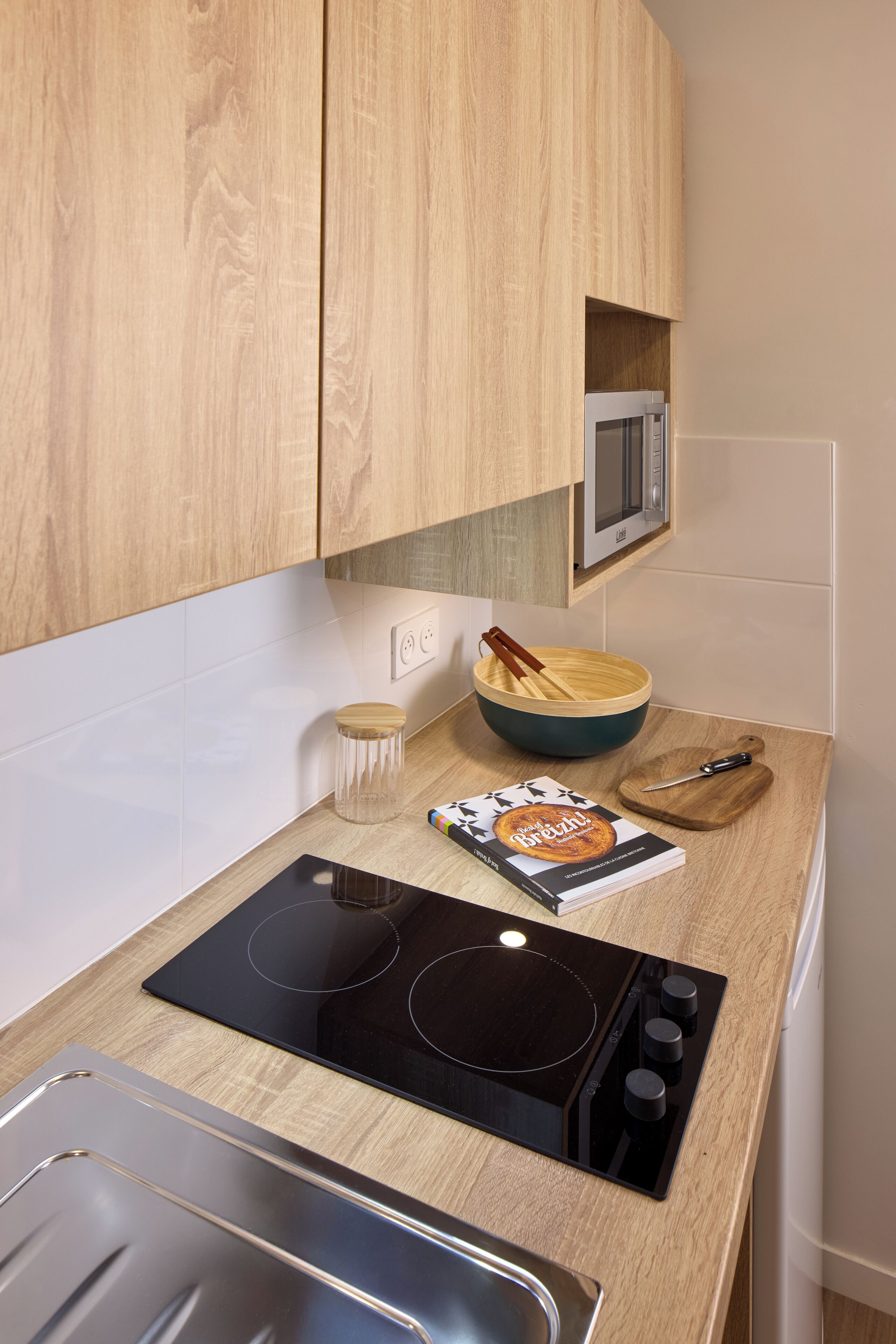 A compact kitchen with wooden cabinets, a black induction cooktop, a microwave, and a bowl with a wooden spoon.