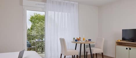 A modern hotel room with a bed, a laptop, a dining table with chairs, a TV, and a balcony with a view of trees.