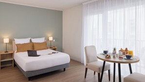 Een hotelkamer met een bed, bedlampen, een eettafel met ontbijtartikelen en een groot raam met dunne gordijnen.