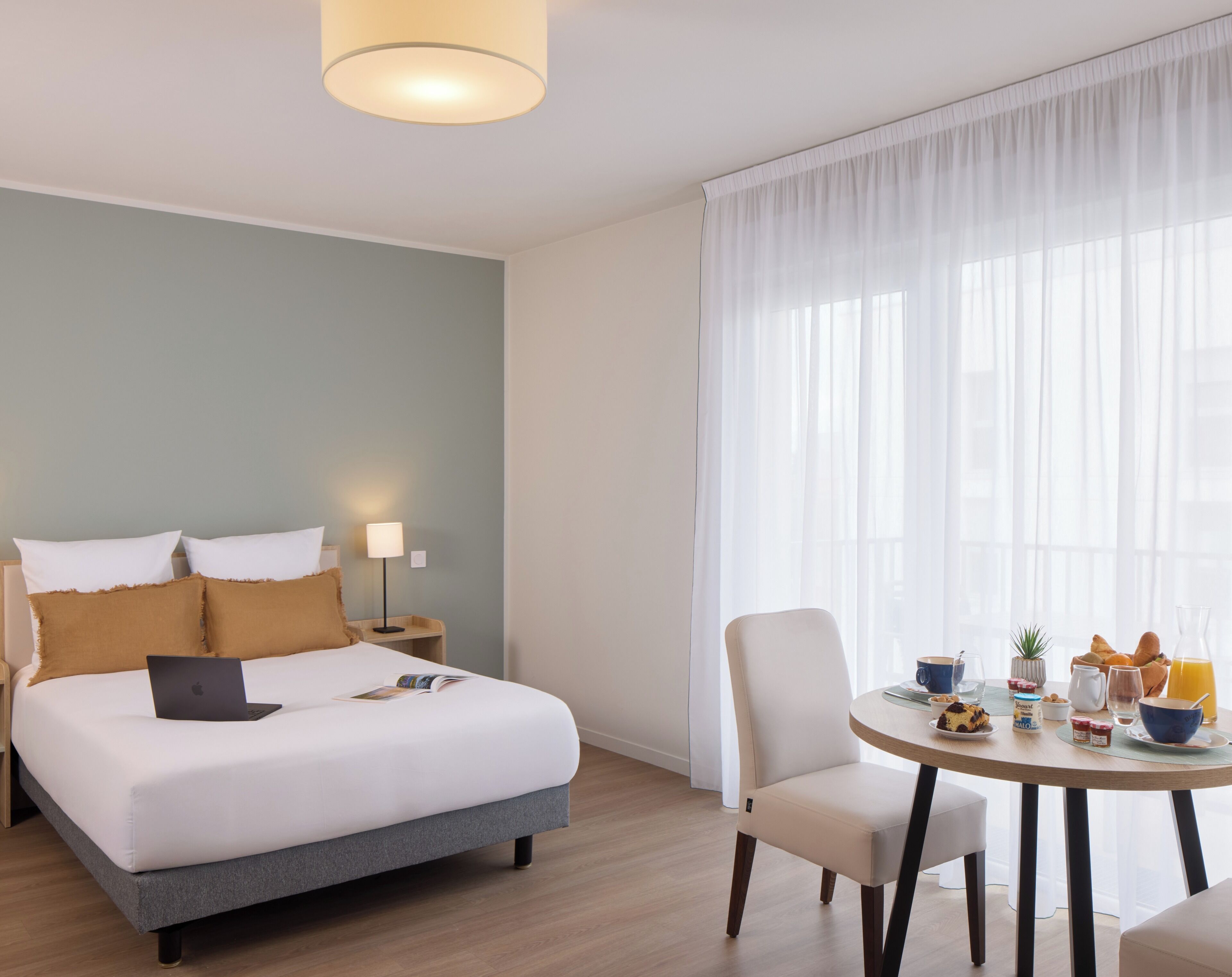 Hotelkamer met een bed, bedlamp, laptop, ontbijttafel met eten en drinken, en een raam met dunne gordijnen.