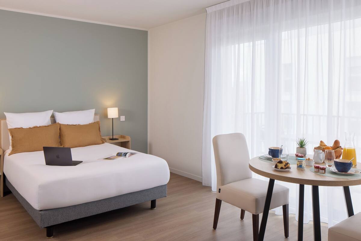 A hotel room with a bed, bedside lamp, laptop, breakfast table with food and drinks, and a window with sheer curtains.