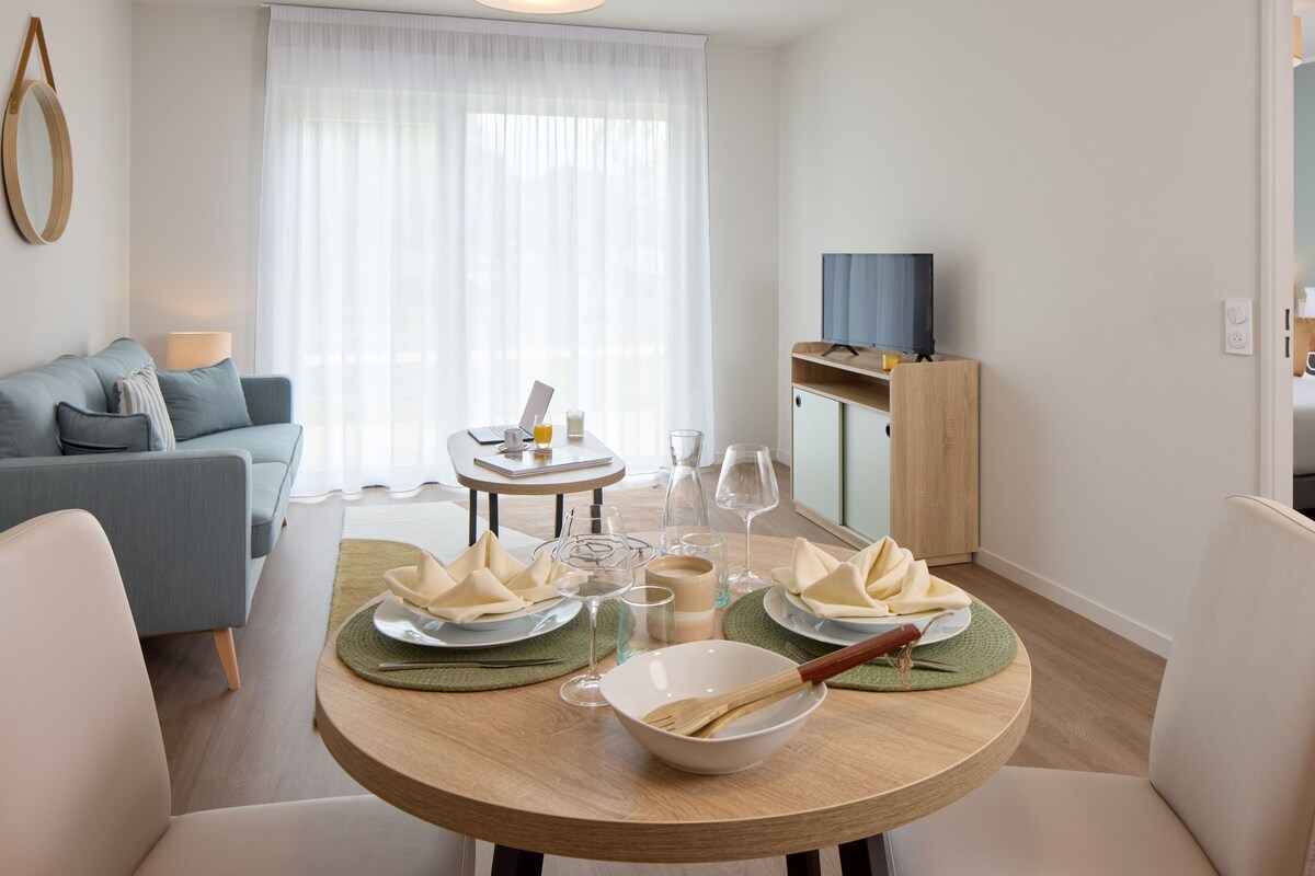 Een moderne woonkamer met een eettafel gedekt voor twee personen, een bank, een televisie en een groot raam met dunne gordijnen.