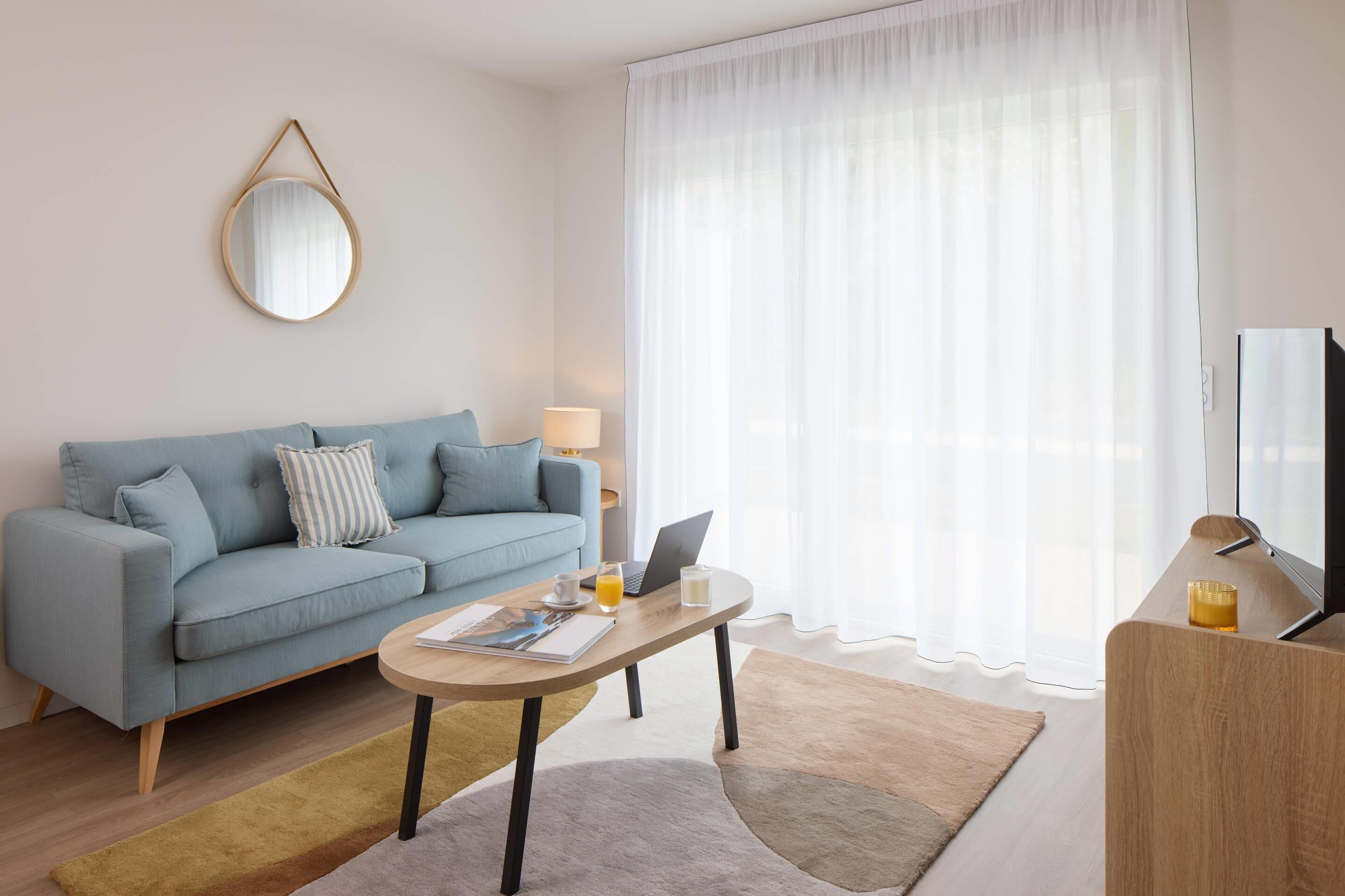 Un salon moderne avec un canapé bleu clair, une table basse ronde et un téléviseur posé sur un meuble en bois.