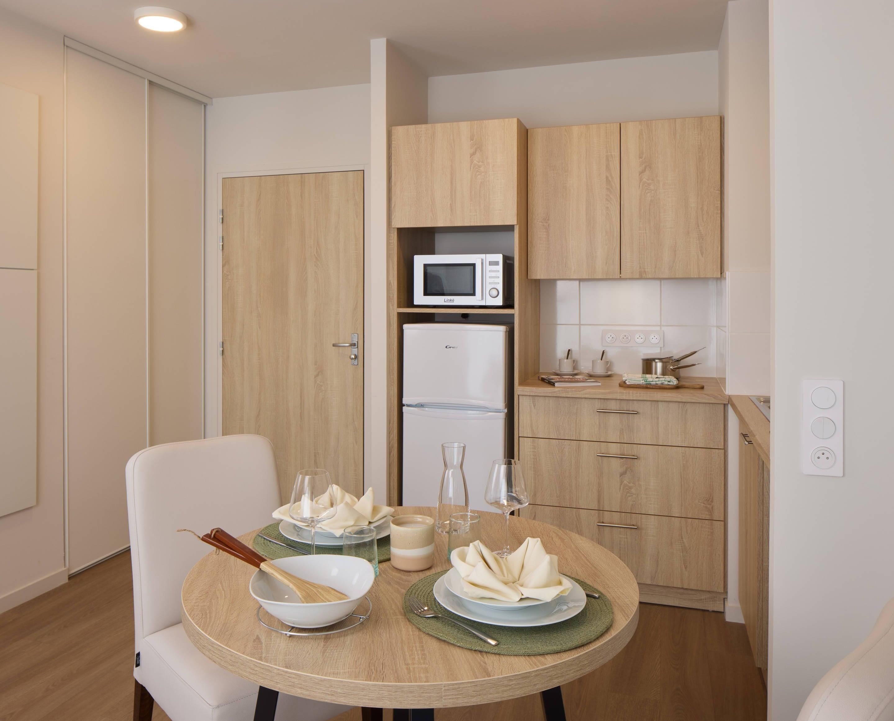 A compact kitchen with a microwave, refrigerator, and wooden cabinets. A dining table with plates, glasses, and a bowl is set for two.