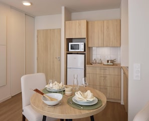 A compact kitchen with a microwave, refrigerator, and wooden cabinets. A dining table with plates, glasses, and a bowl is set for two.