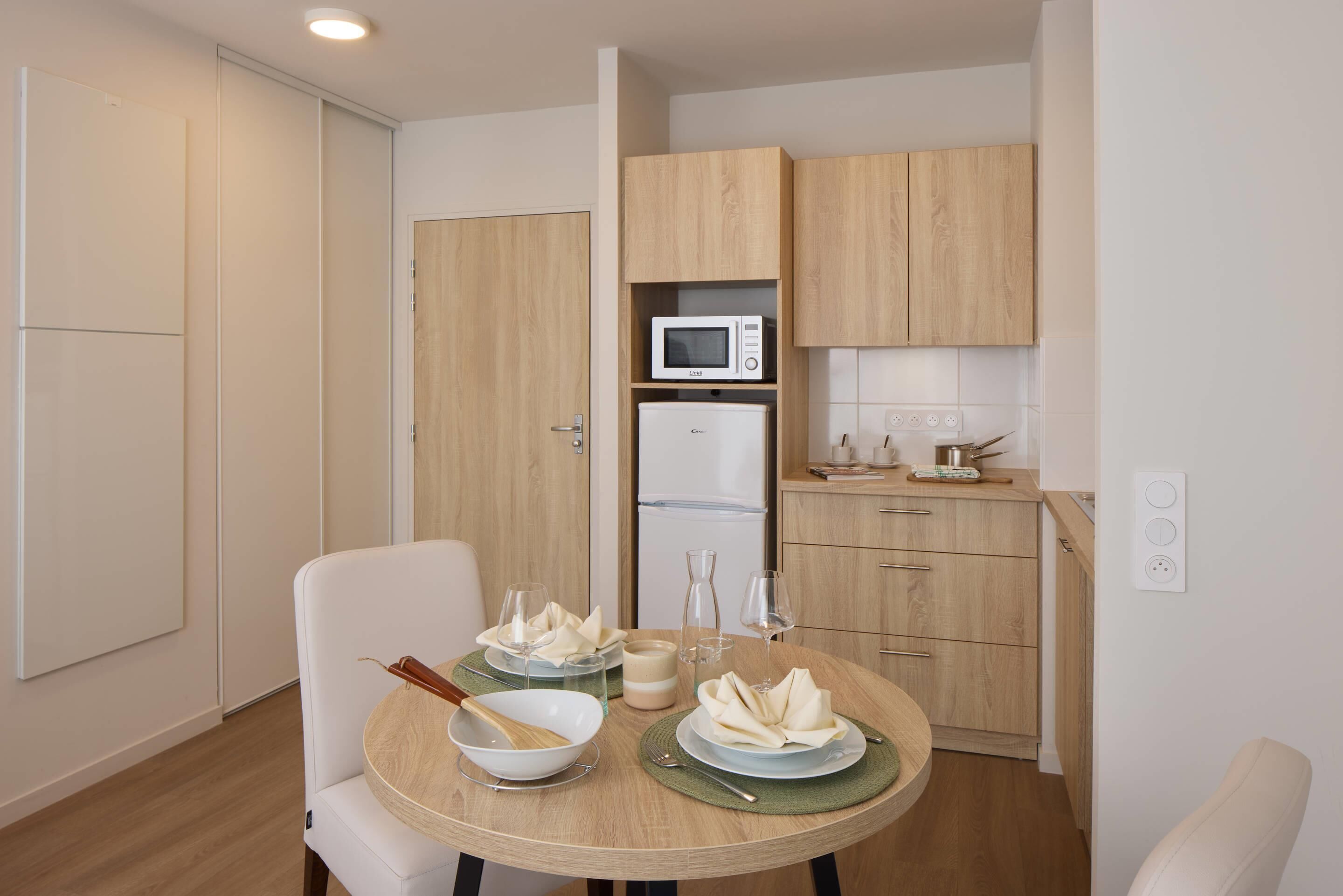 A compact kitchen with a microwave, refrigerator, and wooden cabinets. A dining table with plates and glasses is set up in the room.