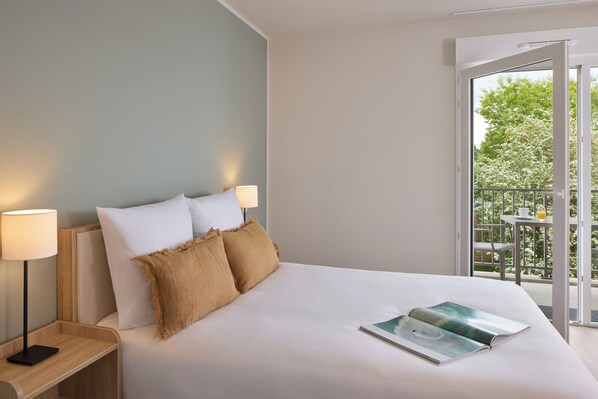 Una habitación de hotel con una cama grande, dos lámparas de noche, una revista sobre la cama y un balcón con mesa y sillas.