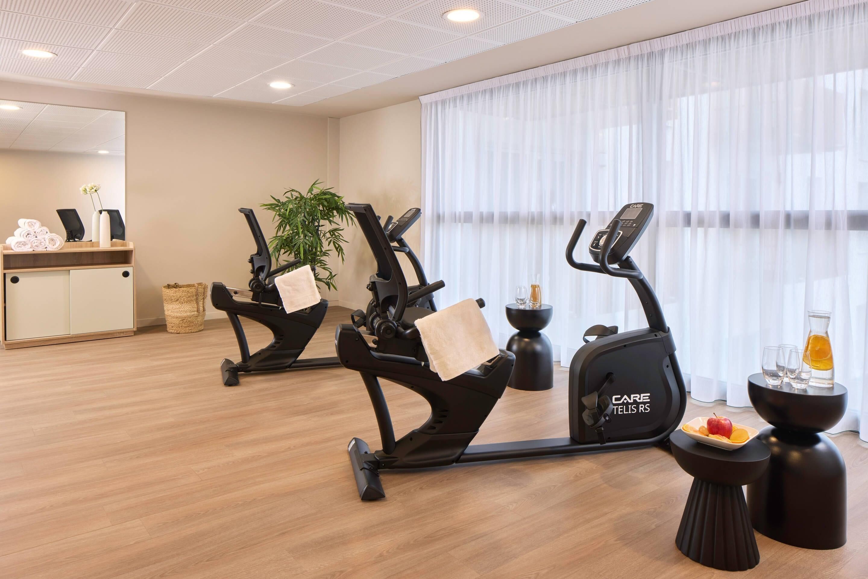 A modern fitness room with two stationary bikes, a small table with a fruit plate, and a large window with sheer curtains.
