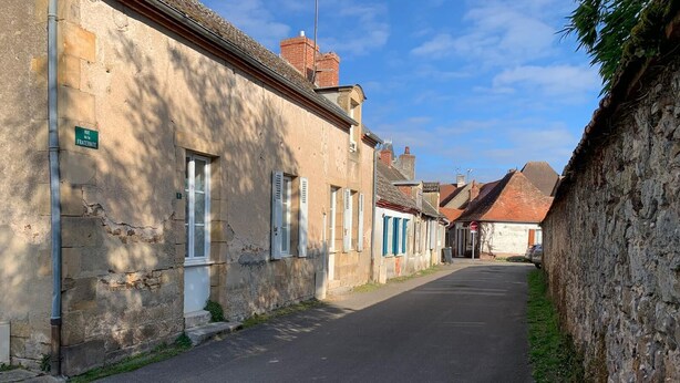 Una calle estrecha bordeada de edificios antiguos de piedra, algunos con contraventanas, bajo un cielo despejado.