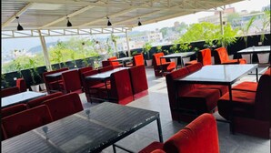 An outdoor restaurant with red seating and tables, featuring a view of buildings and greenery. - Hotel Nikanshvee (Udaipur)