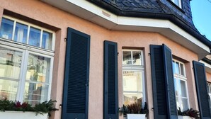 Une maison d’angle avec un toit à forte pente, des bardeaux sombres et une façade en stuc de couleur beige.