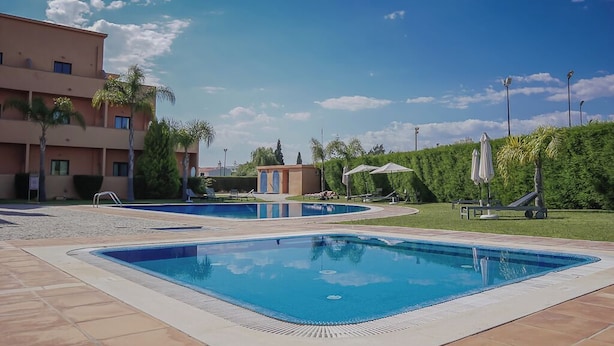 Ein Poolbereich mit zwei Schwimmbecken, einem mehrstöckigen Gebäude und einem gepflegten Rasen mit Palmen.