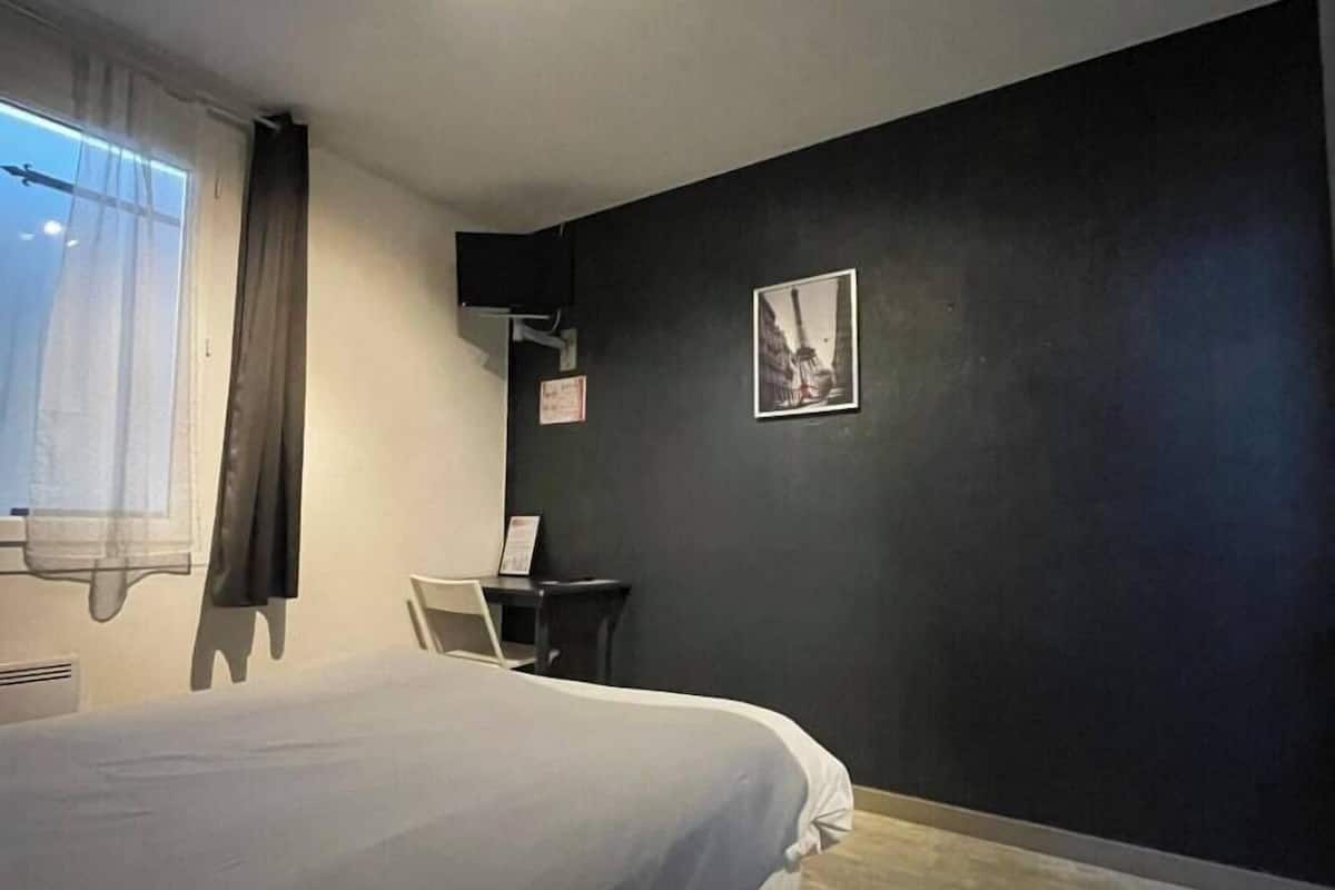 Un dormitorio con cama, escritorio, silla, ventana con cortinas y televisor montado en la pared.
