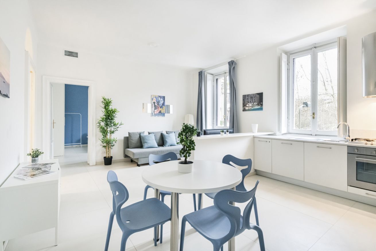 Een moderne keuken met witte kastjes, een eethoek met een ronde tafel en blauwe stoelen, een inbouwoven en een raam met gordijnen.