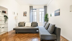 A modern living room with a grey sectional sofa, wooden parquet flooring, a glass shelving unit, and a large window with curtains. - Easylife - Classy apartment in Buenos Aires (Milano)