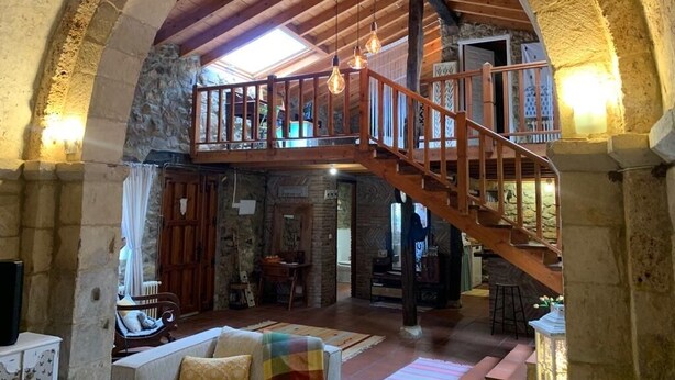Un interior rústico con una escalera de madera que sube a un nivel superior, paredes de piedra y una zona de estar acogedora con cojines.