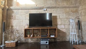Una habitación con pared de piedra, un televisor de pantalla plana sobre un soporte de madera, un altavoz pequeño, una escalera blanca y una cesta de mimbre con ramas.