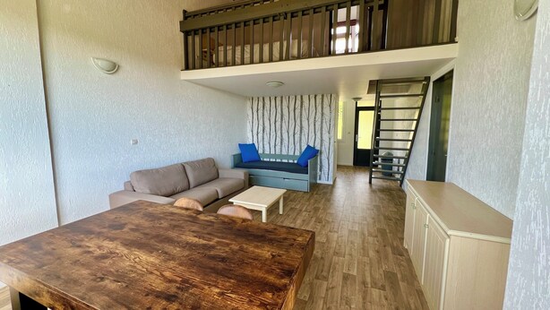 Amplia sala de estar con una mesa de comedor de madera, un sofá y un cojín azul decorativo. Una escalera conduce a un nivel superior con un balcón.