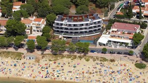 An aerial view of a beach with numerous people and umbrellas, surrounded by residential buildings and a large hotel with multiple floors and balconies.