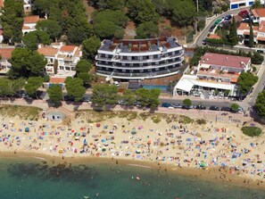 Vista aérea de uma praia com muitas pessoas e guarda-sóis, cercada por edifícios residenciais e um grande hotel com vários andares e varandas.