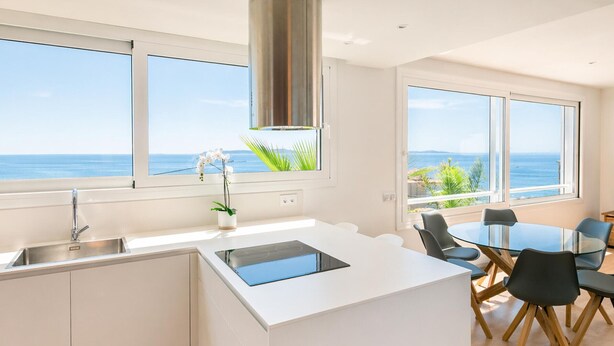 Una cocina moderna con una isla central, un comedor con mesa de vidrio y amplias ventanas con vistas al océano.