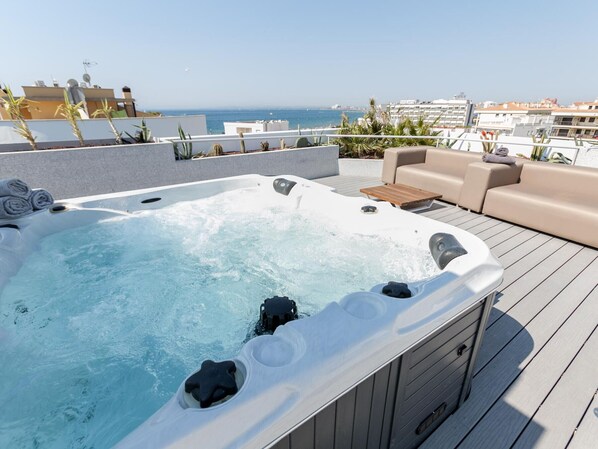 A hot tub on a deck with a view of the sea, a sofa, and a wooden table.