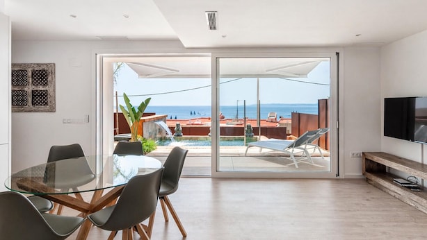 A modern living room with a glass table, chairs, a TV, and a view of a pool and ocean.