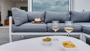 Un salon moderne avec un canapé d’angle gris, une table basse blanche sur laquelle sont posés des en-cas et un verre de vin, et un chapeau sur le canapé.