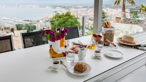 Une table avec des aliments pour le petit-déjeuner, un vase de fleurs et une vue sur une ville côtière.