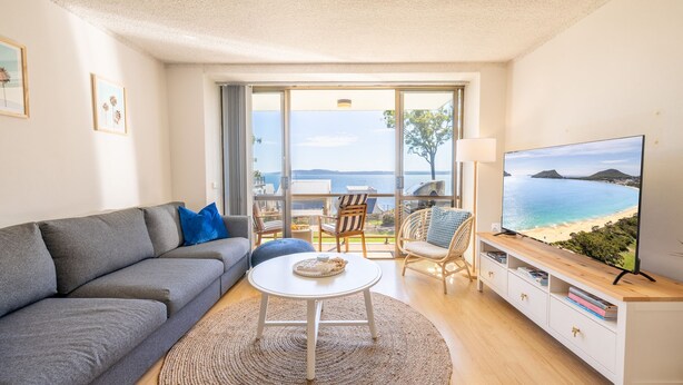 Uma sala de estar moderna com um sofá cinzento, uma mesa redonda branca e uma TV exibindo uma vista de praia.