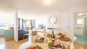 Sala de estar moderna com área de jantar posta para refeição, grandes janelas com vista para o mar e cozinha visível ao fundo.