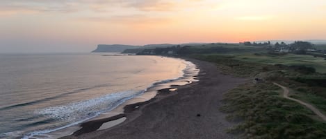 Un paysage côtier avec une plage de sable, un sentier longeant la côte et des collines à l’horizon.