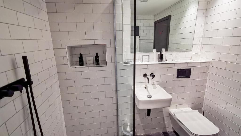 Un baño moderno con paredes de azulejos blancos, cabina de ducha de vidrio, lavamanos blanco y un estante pequeño con artículos de tocador.