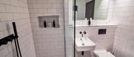 Un baño moderno con paredes de azulejos blancos, cabina de ducha de vidrio, lavamanos blanco y un estante pequeño con artículos de tocador.