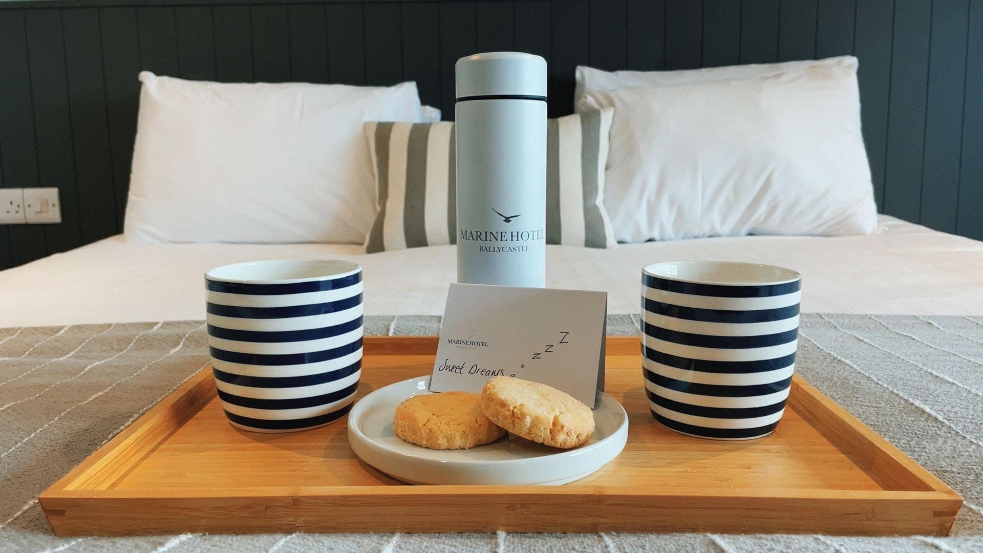 Ein ordentlich angerichteter Frühstückswagen auf einem Bett, mit einer weißen und dunkelblauen Streifen-Tasse, einem Teller mit Keksen, einem Thermosbehälter und einem Zettel.