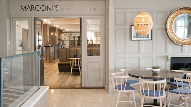 A modern interior of Marconi's Bar & Bistro with a glass partition, a round table with chairs, and a hanging light fixture. - Marine Hotel Ballycastle (Ballycastle)