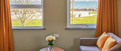A room with two windows overlooking a grassy area and the sea. There is a grey sofa with orange cushions and a small round table with a vase of flowers and a magazine.