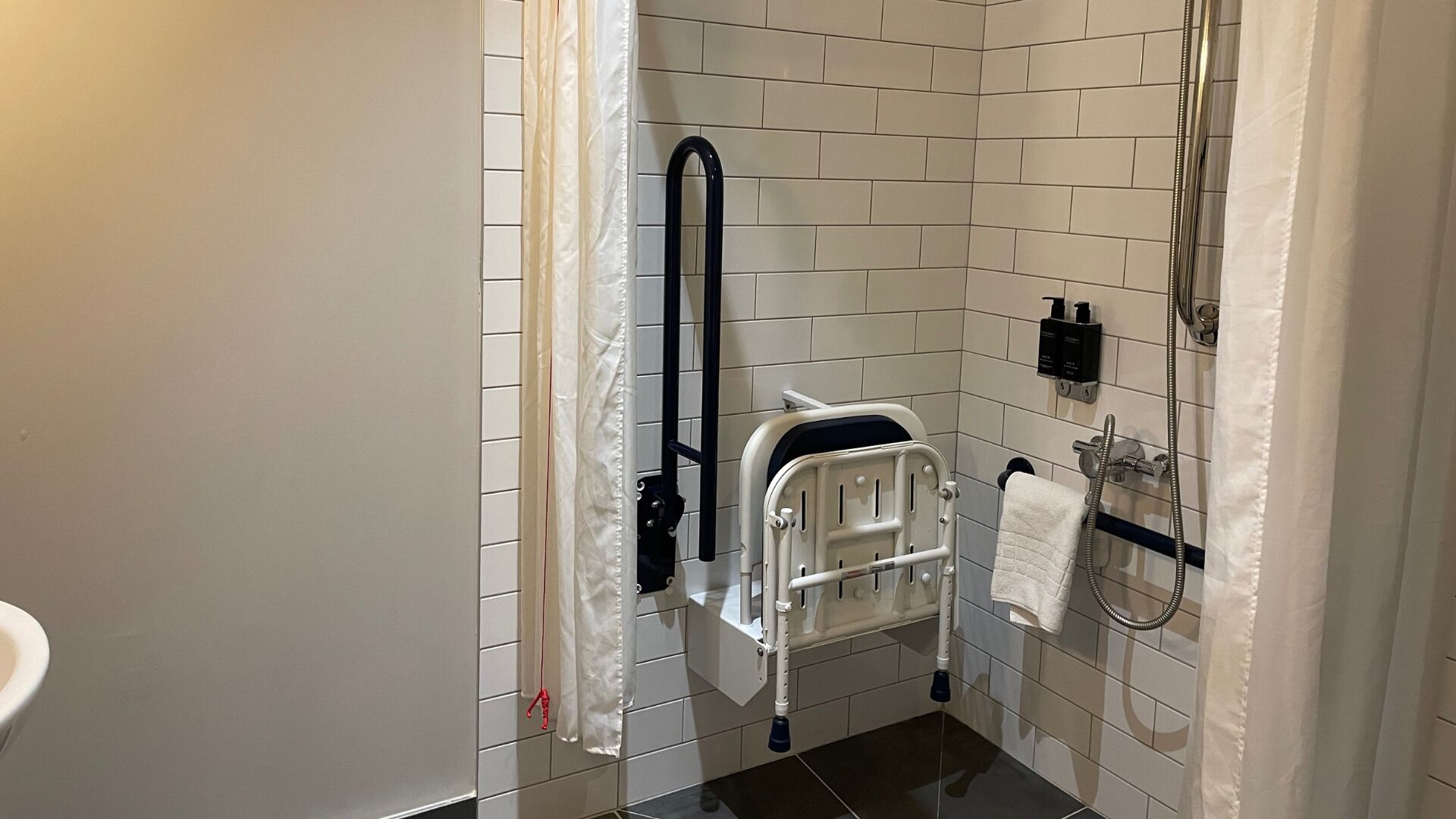 A bathroom with a white tiled floor, a white toilet, a shower with a handheld showerhead, and a white towel hanging on the wall.