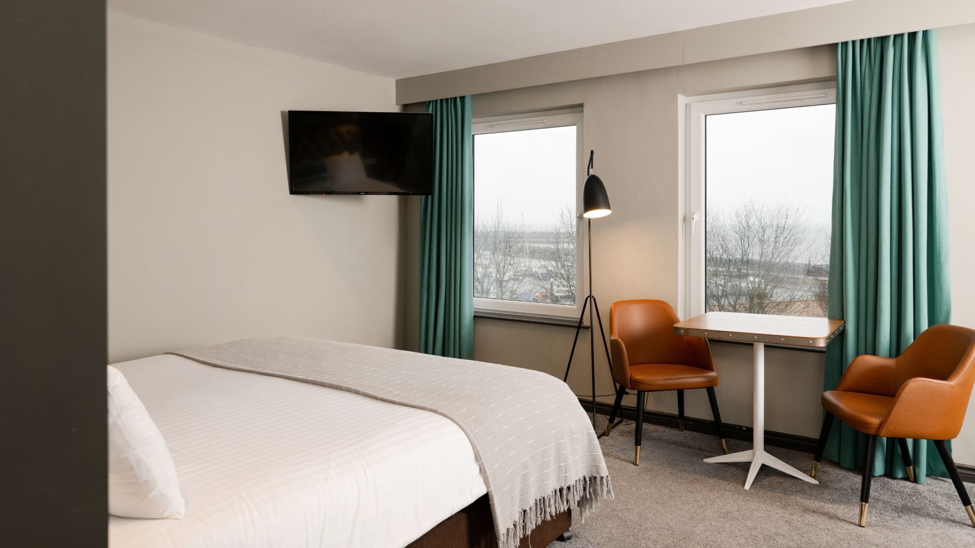 A hotel room with a bed, two chairs, a TV, and a window with curtains.