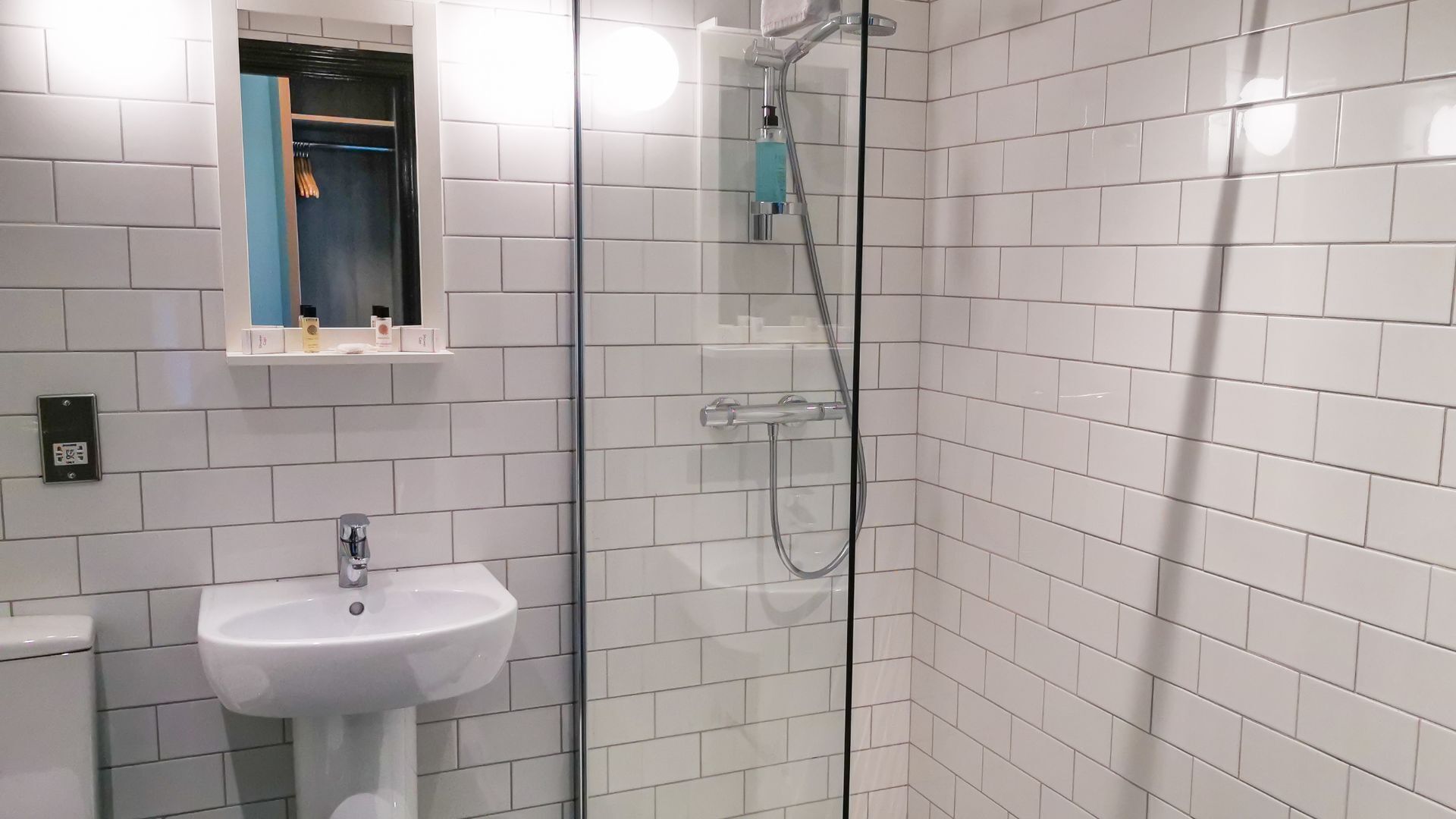 Un baño con paredes de azulejos blancos, un lavamanos blanco y una zona de ducha con mampara de vidrio.