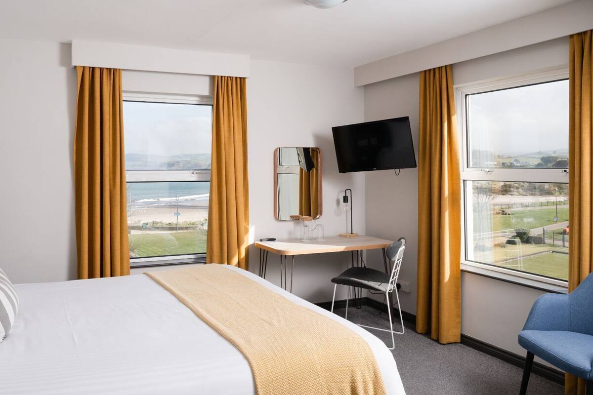 Ein Hotelzimmer mit Bett, Schreibtisch, Stuhl, Fernseher und großen Fenstern mit Vorhängen.