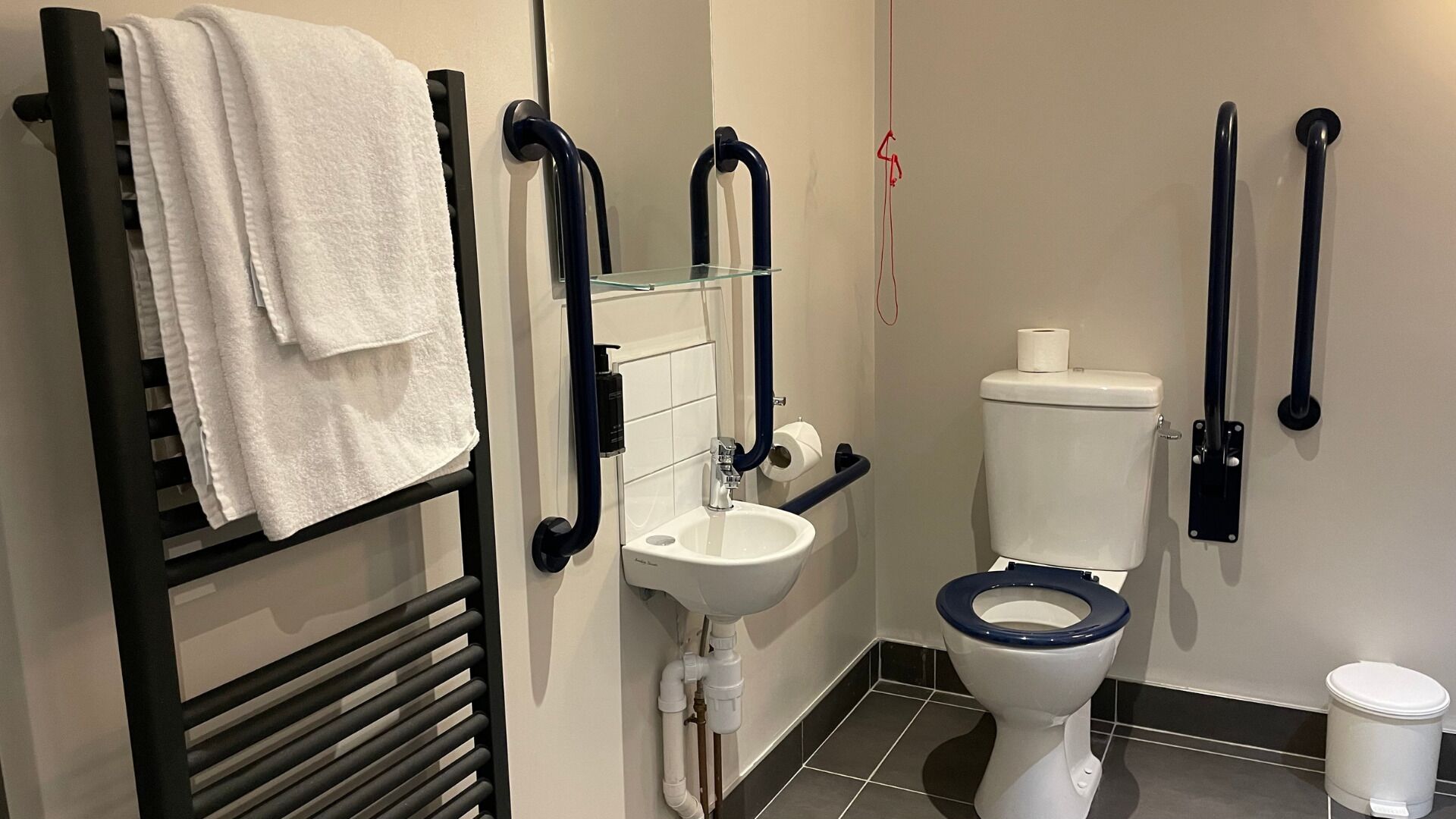 A modern bathroom with a black towel rack, a glass shelf, a white sink, a toilet with a blue seat, and a white trash bin.